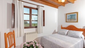 Een slaapkamer met een houten hoofdbord, een bed met beddengoed met een patroon, een raam met gordijnen, een radiator en een lijst met een schilderij aan de muur.