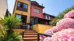 A two-story house with a red and yellow facade, a visible staircase, and lush greenery including pink hydrangeas.