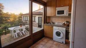 A compact kitchen with a washing machine, a microwave, and a sink. There is a balcony with a table and chairs, and a view of a building with a tiled roof.