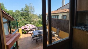 A patio with a table and chairs, a view of a house with a tiled roof, and a garden with pink flowers.
