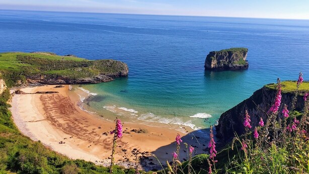 A coastal landscape with a sandy beach, rocky cliffs, and clear blue sea.