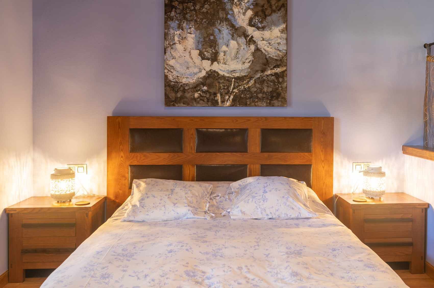 Ein Schlafzimmer mit einem Holz-Kopfteil, einem blumigen Bettbezug, zwei Nachttischlampen und einem Bild an der Wand.