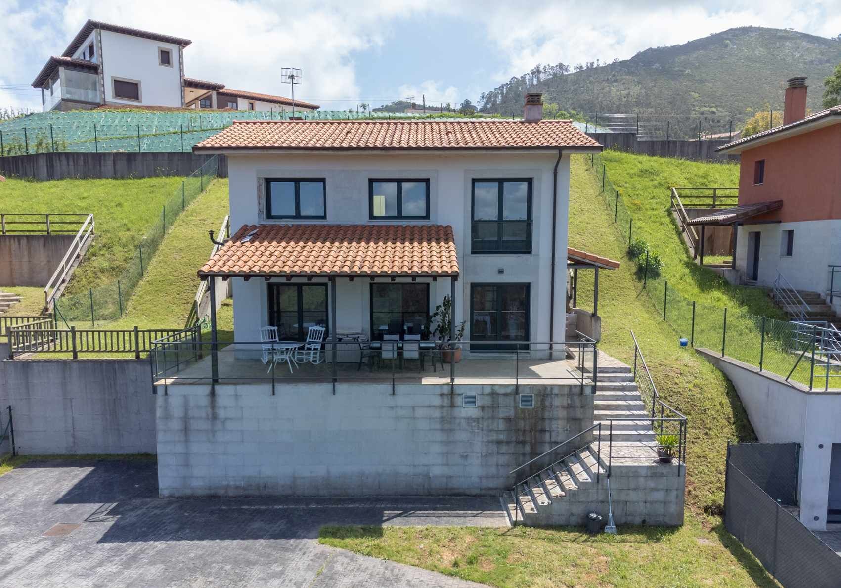 Une maison à deux étages, avec un toit recouvert de tuiles, un patio couvert aménagé avec du mobilier d’extérieur et un escalier en béton menant à l’entrée.