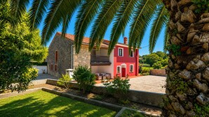 A two-story house with a stone facade and a red upper section, surrounded by a well-maintained garden and palm trees. - Casa La Venta de Pedro (Ribadesella)