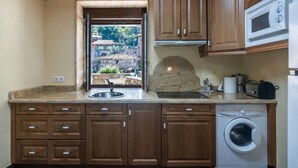 Cozinha com armários de madeira, máquina de lavar embutida, micro-ondas e janela com vista para uma casa e vegetação.