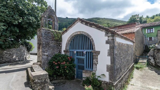 Um pequeno edifício de pedra com porta azul, rodeado por vegetação e um muro de pedra.