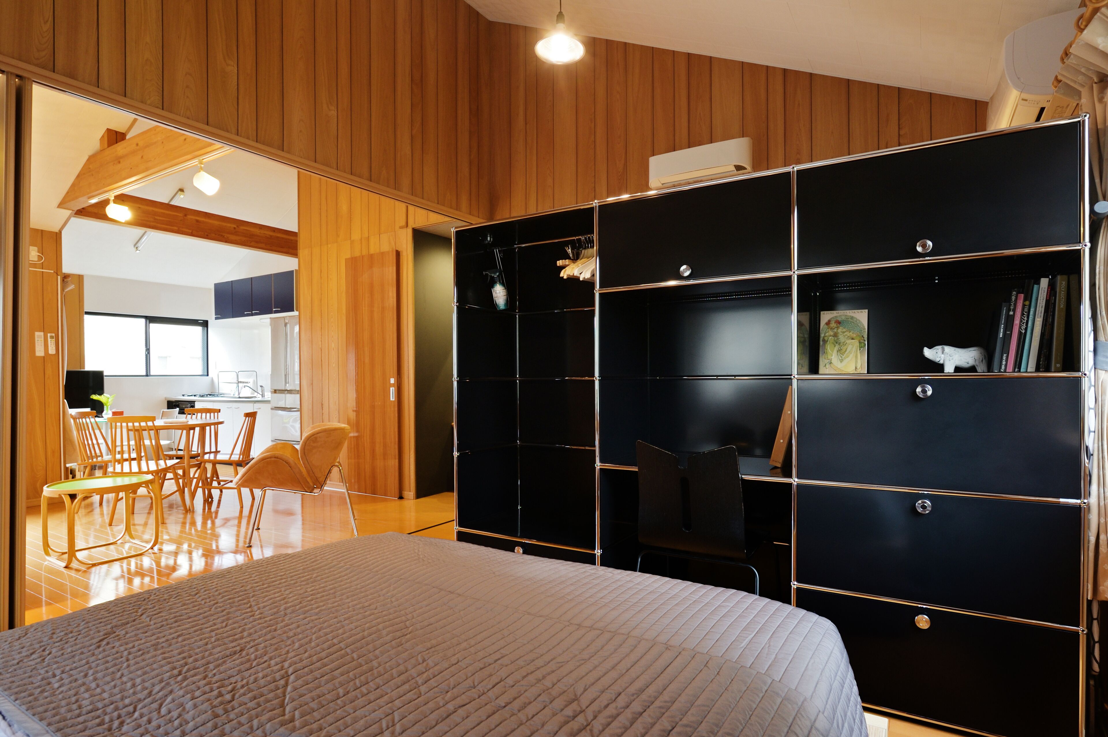 A modern bedroom with a large bed, wooden panel walls, a black wardrobe, and a dining area with chairs and a table.