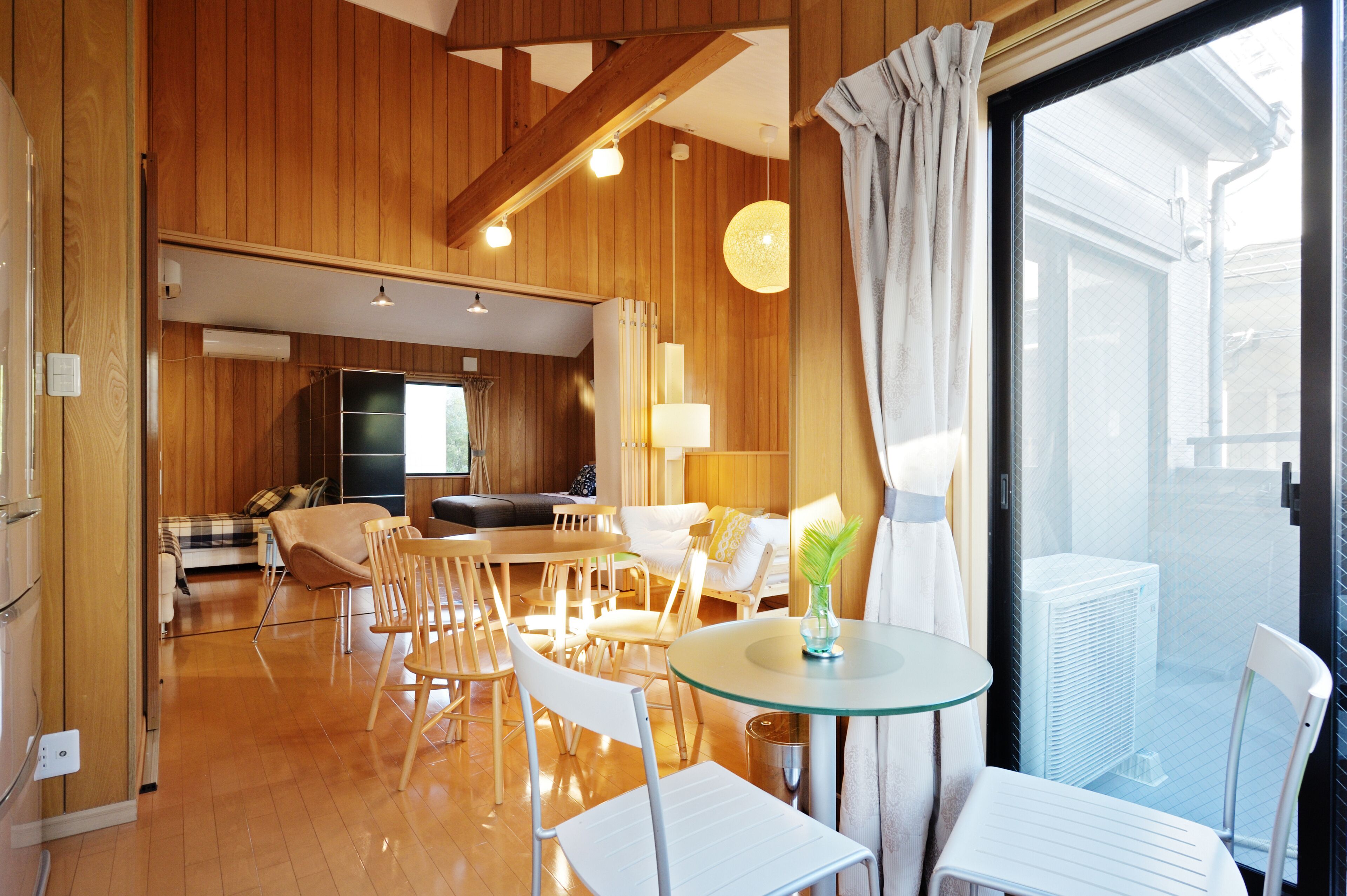 Een modern interieur met houten lambrisering, een eetgedeelte met witte stoelen, een glazen tafel en een groot raam met gordijnen.