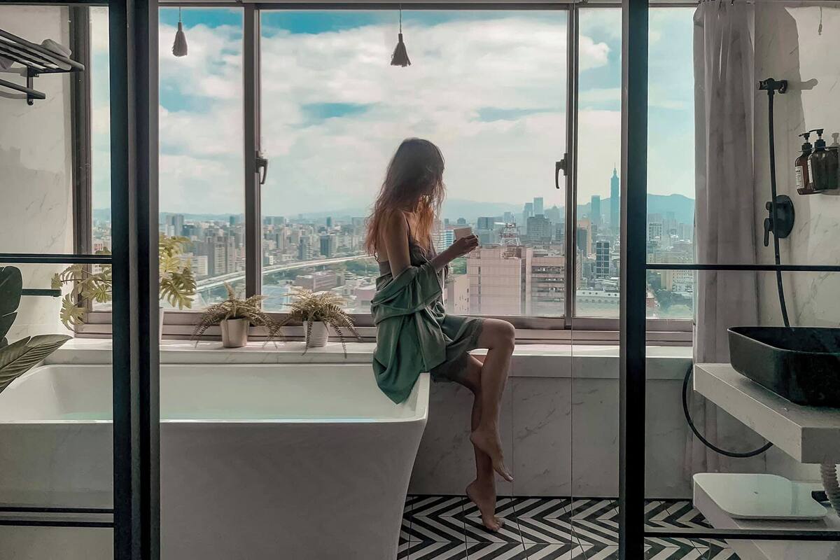 A modern bathroom with a large bathtub, a view of the cityscape, and a potted plant on the windowsill.