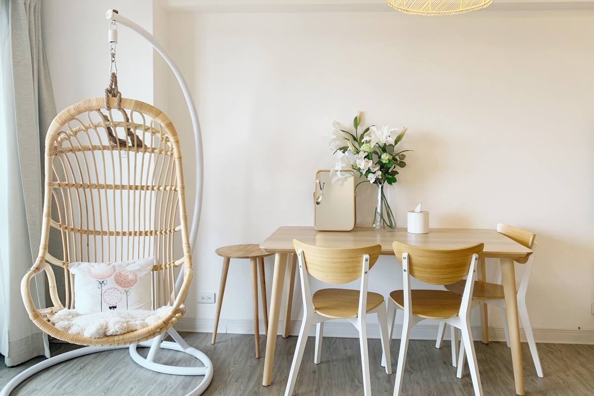 A dining area with a wooden table, white chairs, a hanging chair, a vase with flowers, and a lamp.