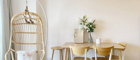 A dining area with a wooden table, white chairs, a hanging chair, a vase with flowers, and a lamp.