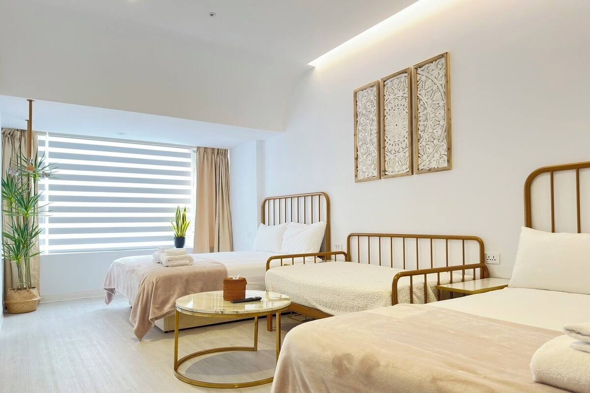 A hotel room with two single beds, a round table, a potted plant, and a window with blinds.
