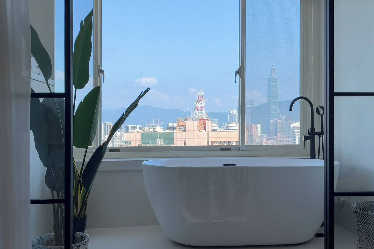 A modern bathroom with a freestanding bathtub, a window with a cityscape view, and a potted plant.