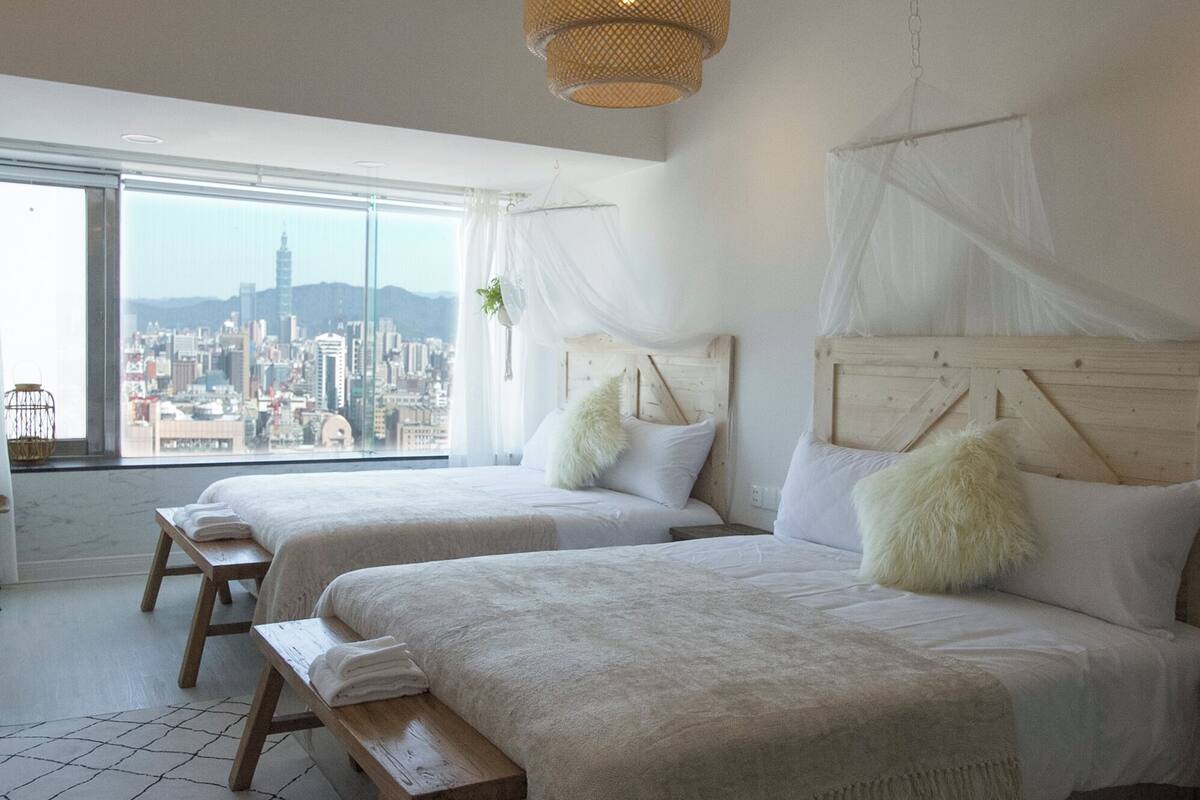 A hotel room with two beds, a large window with a city view, and a hanging plant.