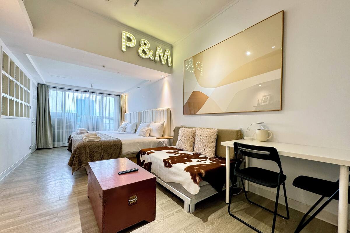 A hotel room with a bed, a desk, a chair, a small table, and a large window.