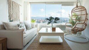 A modern living room with a white sofa, a coffee table, a wicker chair, and a large window offering a city view. - Urban Abode Apartment 1 (Taipei)