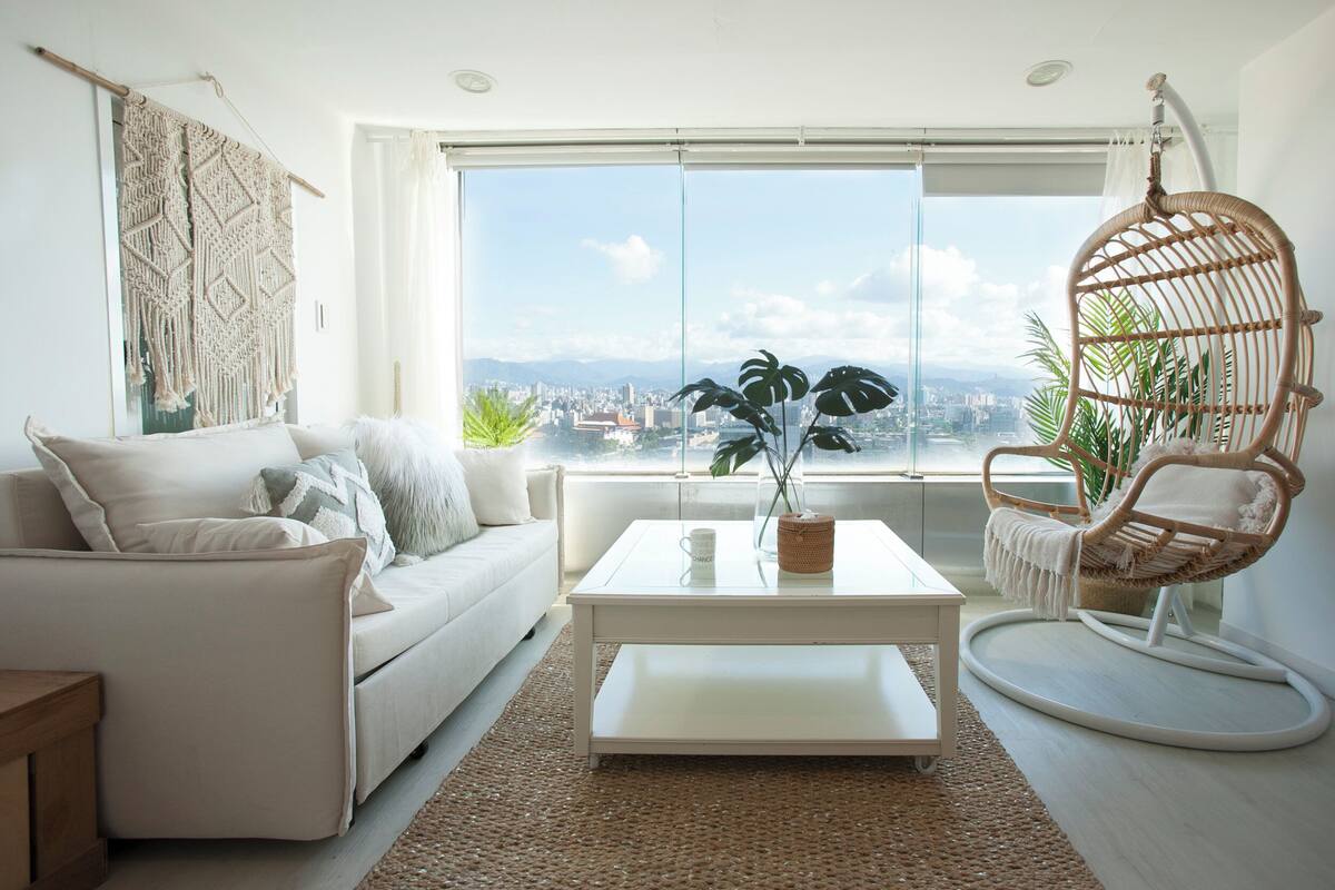 A modern living room with a white sofa, a coffee table, a wicker chair, and a large window offering a city view.