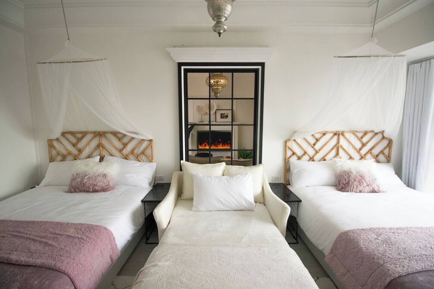 Habitación con dos camas, cada una con cabecero de madera, ropa de cama blanca y una manta rosa. Se ve un fuego a través de la ventana, y un candelabro cuelga del techo.
