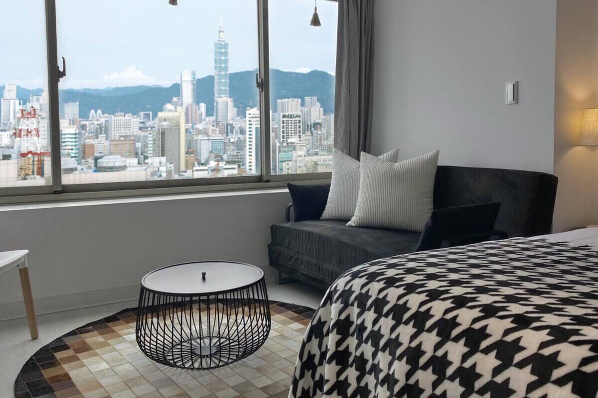 A hotel room with a large window offering a city view, a round table, a bed with patterned bedding, and a grey sofa with cushions.