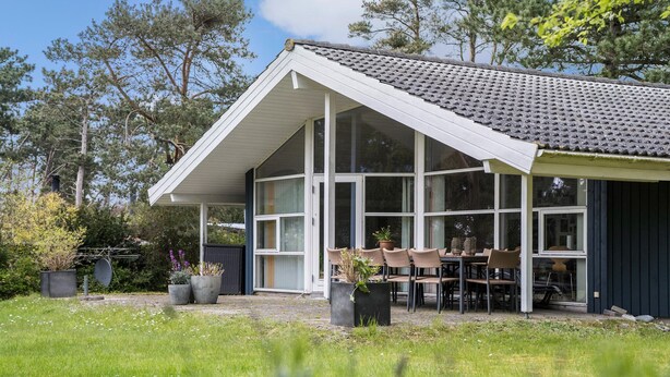 Une maison dotée d’une grande baie vitrée, d’une table à manger avec des chaises et d’un jardin agrémenté de plantes en pot.