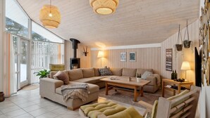 Un salon chaleureux avec un canapé d’angle, une table basse et une cheminée. Des luminaires suspendus et une grande fenêtre laissent entrer la lumière naturelle.