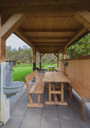 Un espace extérieur abrité, aménagé pour les repas, avec des bancs et des tables en bois, une fontaine et une vue sur un jardin verdoyant.
