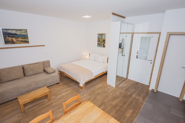 A bedroom with a bed, a sofa, a wooden table, and a chair. There is a door with a mirror and a window.