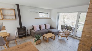 A modern living room with a sofa, coffee table, and a fireplace. There is a dining area with a table and chairs, and a large window providing a view of the outdoors. - Panoramic apartment with Terrace (Visoko)