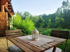 Ein Holztisch mit einer Laterne und einer Unterlage, umgeben von zwei Holzstühlen, auf einer Terrasse mit Blick auf einen üppigen Garten und Bäume.