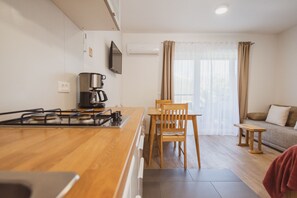 A modern kitchen with a wooden countertop, a gas stove, and a coffee maker. There is a dining area with a wooden table and chairs. A sofa and a small bench are visible in the background. - Classic Studio with Patio (Visoko)