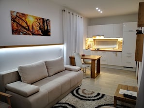 A modern living room with a beige sofa, a dining table, and a kitchen area with white cabinets and a wooden countertop. - Classic Studio with Patio (Visoko)