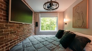 Ett rum med en tegelvägg, en säng med ett quiltat överkast, ett fönster med persienner, en väggmonterad lampa och en takfläkt.