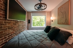 A room with a brick wall, a bed with a quilted bedspread, a window with blinds, a wall-mounted light, and a ceiling fan.