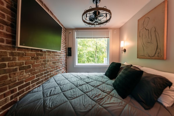 A room with a brick wall, a bed with a quilted bedspread, a window with blinds, a wall-mounted light, and a ceiling fan.