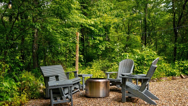 En utomhusplats med sittmöbler, inklusive Adirondack-stolar, och en eldstad av metall, omgiven av grus, belägen i ett skogsområde.