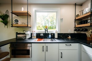 A modern kitchen with a black countertop, stainless steel appliances, and a window with a view of trees. - The Juice Box | Hocking Hills, Container Cabin (Creola)