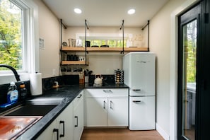 A modern kitchen with white cabinets, a black countertop, a white refrigerator, and a window with a view of trees. - The Juice Box | Hocking Hills, Container Cabin (Creola)