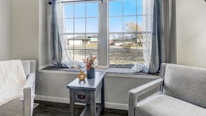 A room with a large window, a small table with a vase, and two chairs. - Pueblo Place (Pueblo)