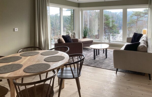 A modern living room with a wooden dining table, two sofas, and a view of a forest through large windows.