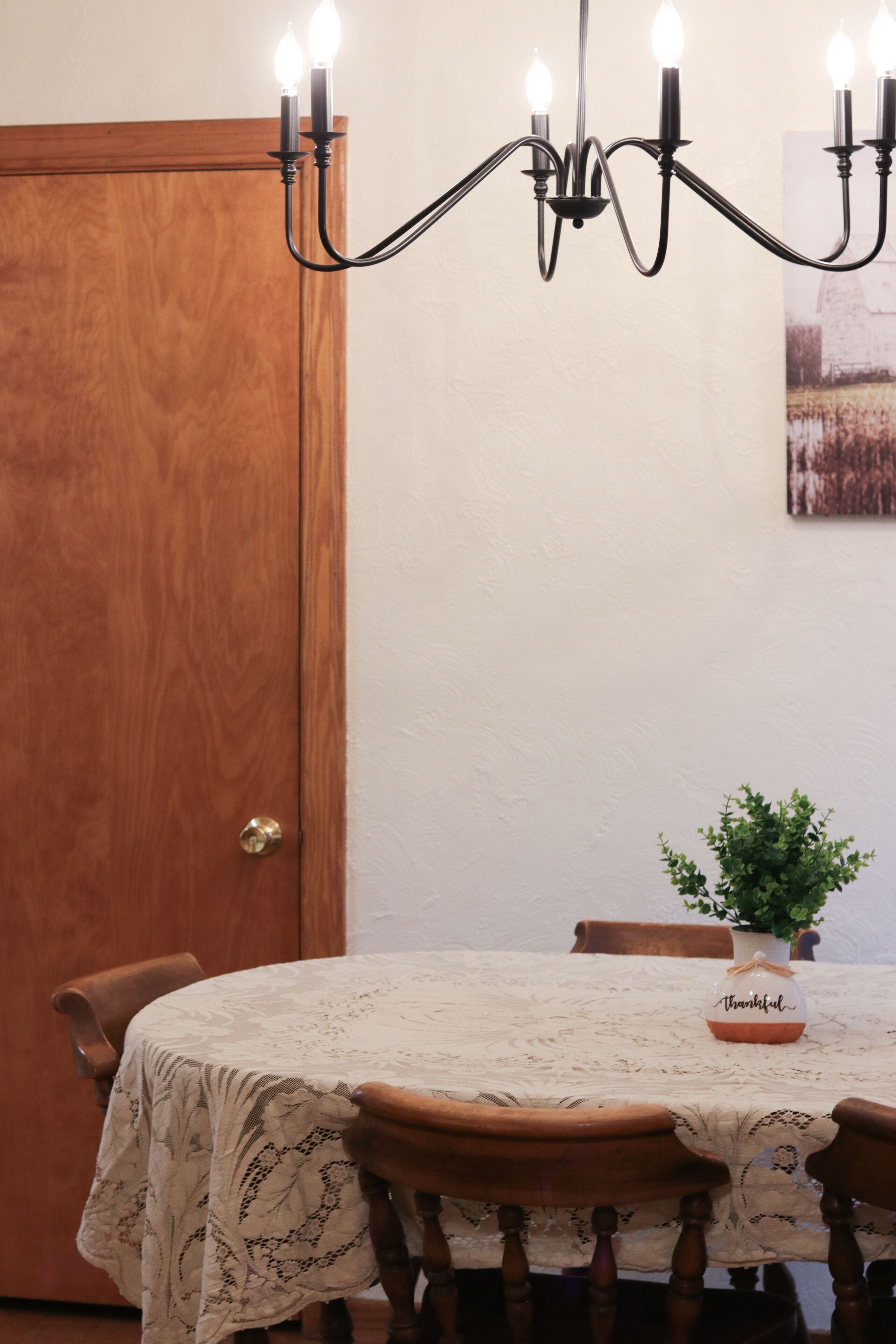 A dining area with a chandelier, wooden chairs, a table with a lace tablecloth, and a potted plant.