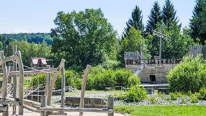 Un parque infantil con estructuras de madera y un tobogán, rodeado de vegetación y un edificio con fachada de madera.