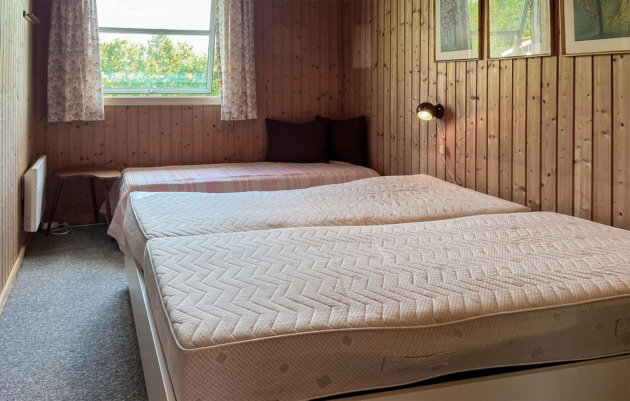 Ein Zimmer mit zwei Betten, Holzwänden, einem Fenster mit Vorhängen, einem Heizkörper und gerahmten Bildern an der Wand.