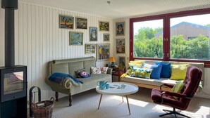 En hyggelig stue med sofa, lænestol og et lille bord. Væggen er prydet med billeder, og fra vinduet kan man se grønne omgivelser.