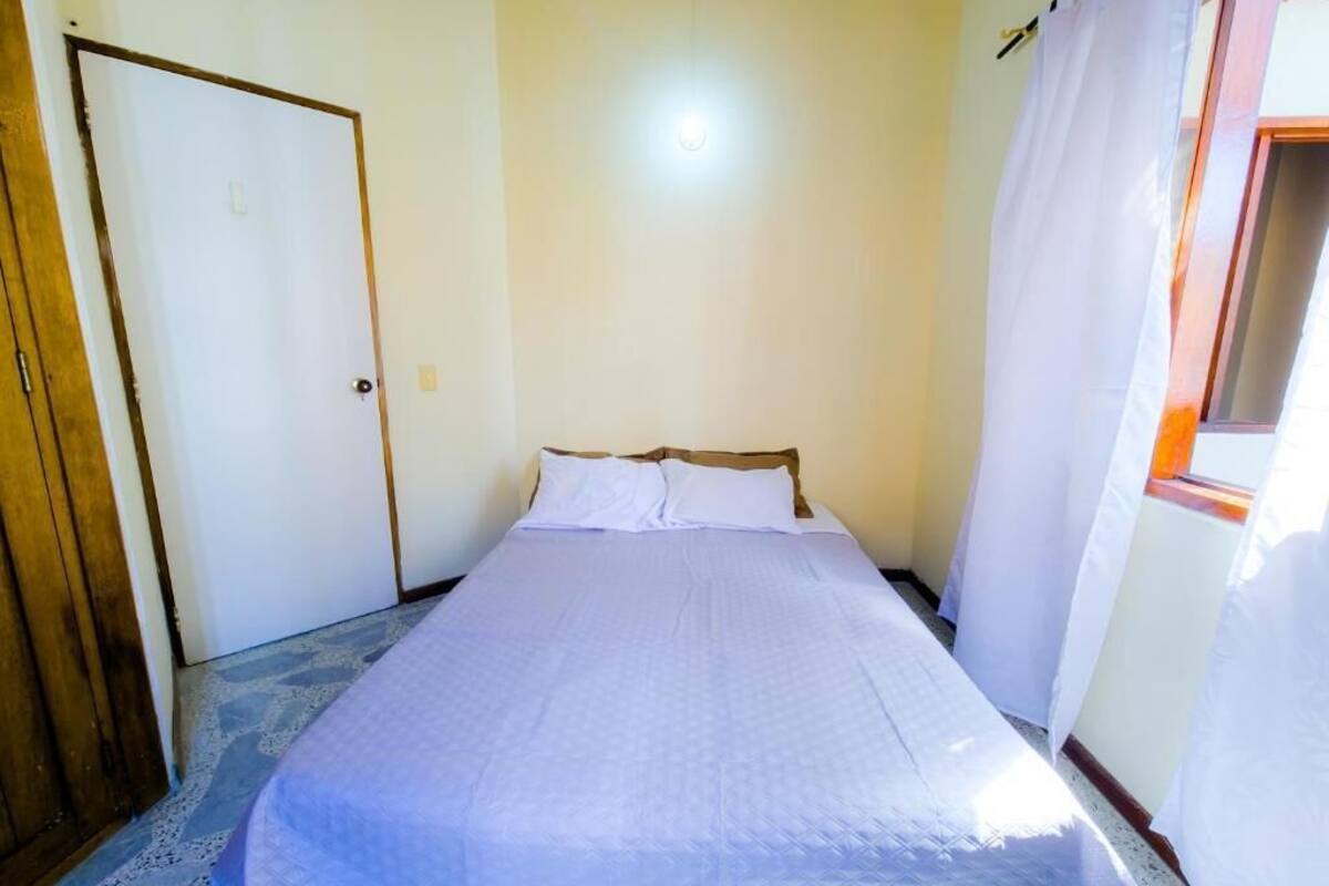 A single-bed room with a wooden door, a window with curtains, and a plain wall.