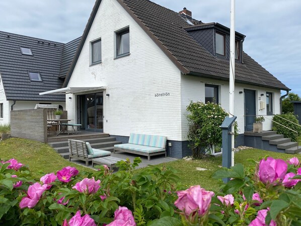 A white two-story house with a sloped roof, a covered porch with a table and chairs, and a small garden with pink flowers.