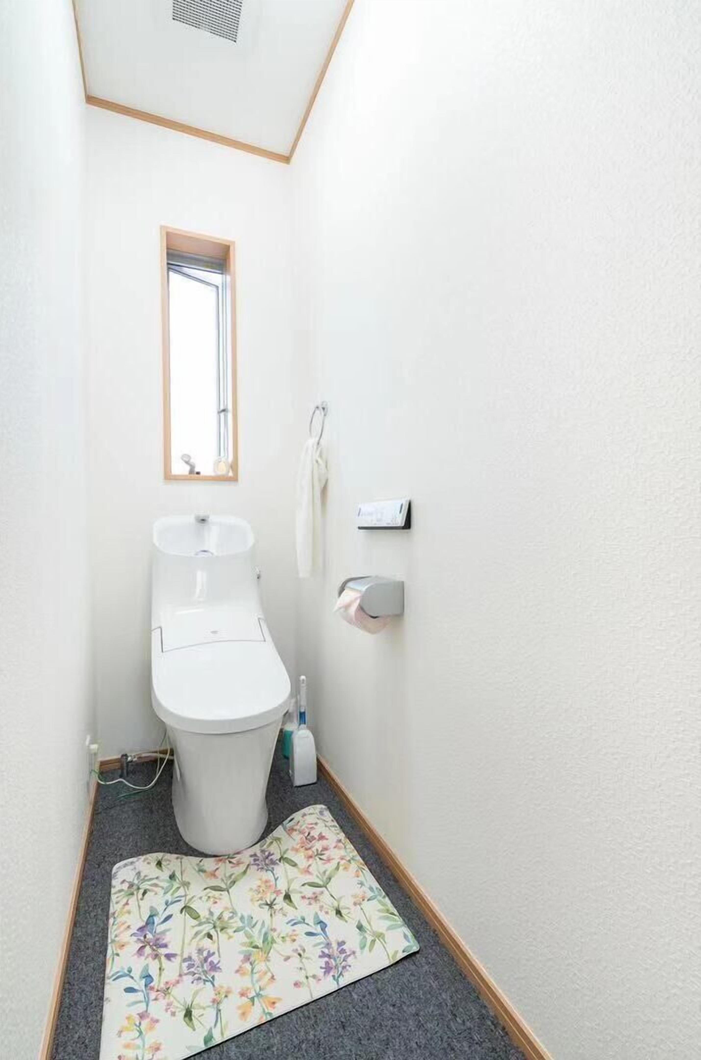 A narrow bathroom with a toilet, a small window, and a towel dispenser.