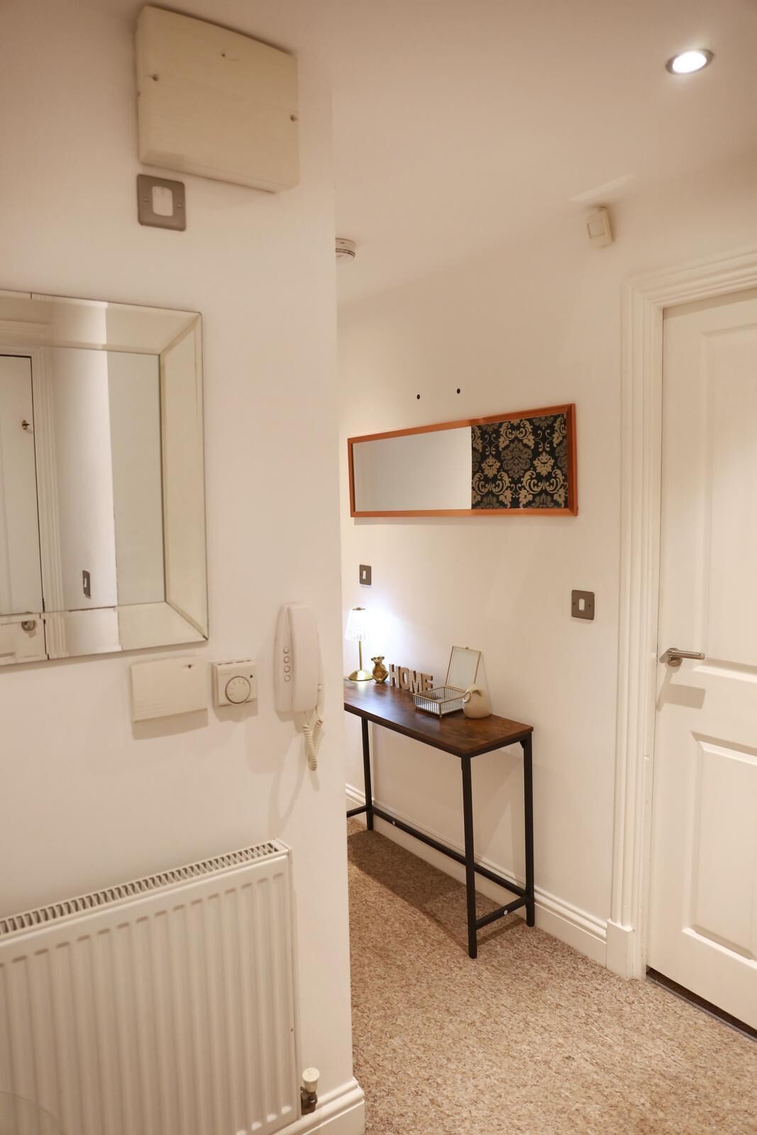 A hallway with a radiator, a telephone, a mirror, a decorative mirror frame, a small table with a lamp, and a door.