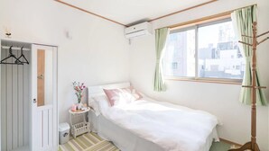 A small, neatly arranged bedroom with a bed, a bedside table with a vase of flowers, a wardrobe, and a window with curtains. - 1 bedroom apartment in saitama (川口市西川口)