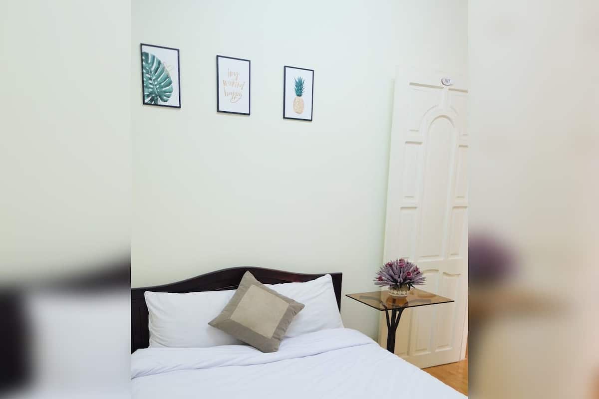 Una cama bien hecha con sábanas blancas y una manta color beige, una mesita de noche con un florero y tres cuadros en la pared.