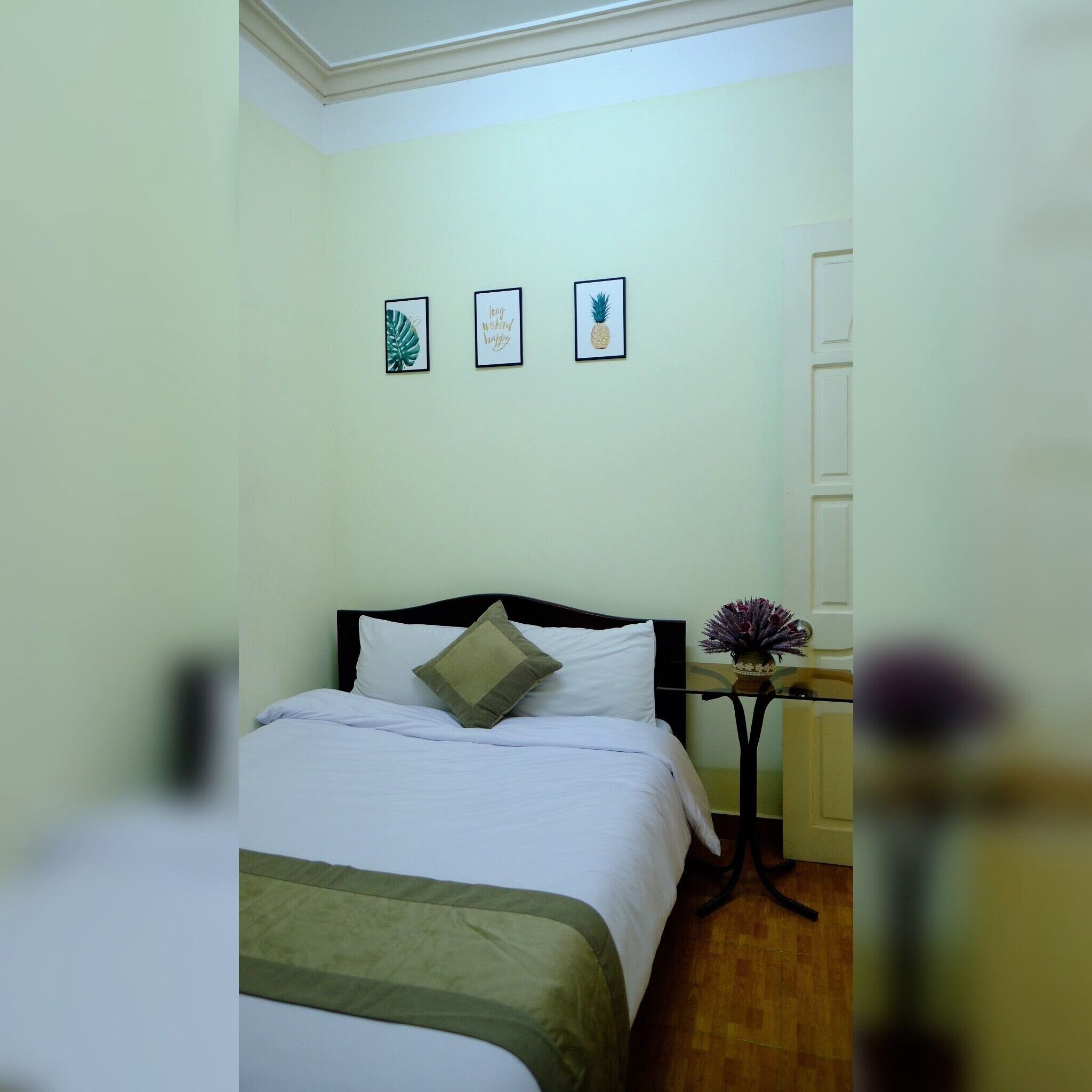A neatly made bed with a green throw, a small bedside table with a vase, and three framed pictures on the wall.